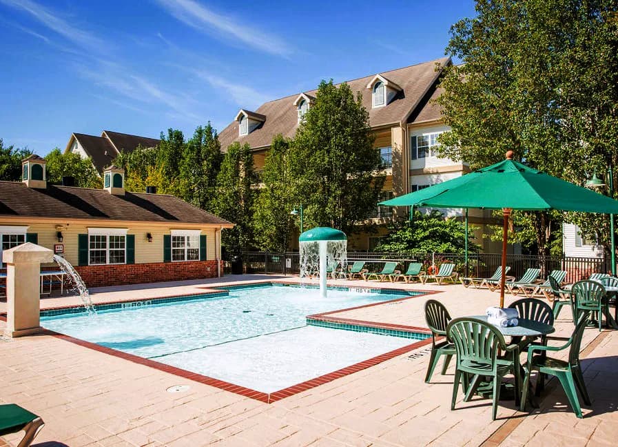 Outdoor pool area with shallow water features, lounge chairs, and green umbrellas beside a clubhouse surrounded by tall trees on a bright sunny day.