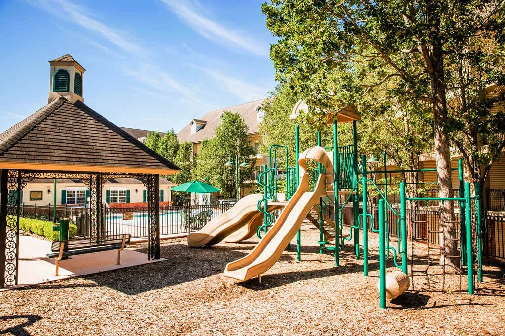 Playground with tan slides, green climbing structures, and a shaded gazebo beside a fenced outdoor pool area surrounded by trees and sunny skies.