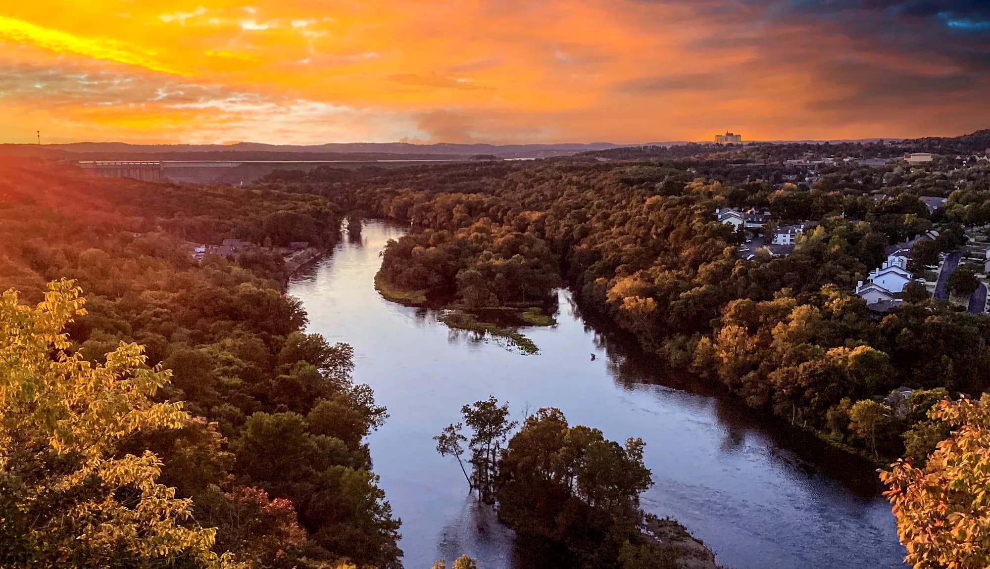 Golden sunset over a winding river surrounded by dense green forest and small town homes, with the sky glowing in orange and purple hues.